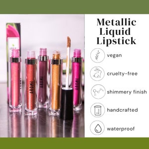 K&ouml;nnte beinhalten: F&uuml;nf Tuben mit metallischem fl&uuml;ssigem Lippenstift in verschiedenen Farbt&ouml;nen von Pink, Gold und Rot. Die Tuben sind mit "AROMI" beschriftet und der Lippenstift wird als vegan, tierversuchsfrei, schimmernd, handgefertigt und wasserfest beschrieben.