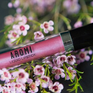 May include: A pink liquid lipstick tube with the brand name "AROMI" and the text "LIQUID LIPSTICK" printed on the label. The tube is surrounded by pink flowers.