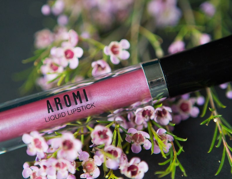 May include: A pink liquid lipstick tube with the brand name "AROMI" and the text "LIQUID LIPSTICK" printed on the label. The tube is surrounded by pink flowers.