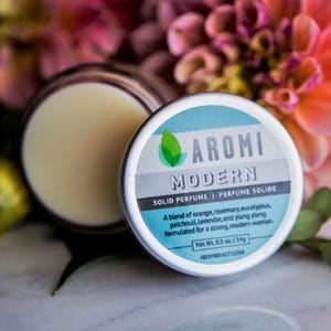 May include: A small, silver tin with a blue and white label that reads "AROMI MODERN SOLID PERFUME | PERFUME SOLIDE". The label also includes a description of the perfume's scent and ingredients, as well as the net weight (0.5 oz. / 14g) and the website address, "AROMIBEAUTY.COM".