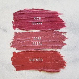 May include: Three swatches of lipstick on a white surface. The swatches are labeled "Rich Berry", "Rose Petal", and "Nutmeg".