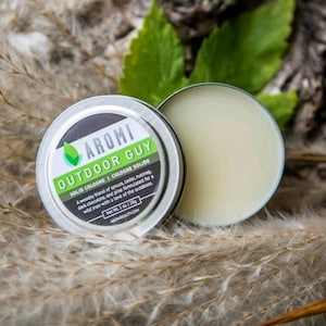 Op de afbeelding: Een zilveren blik met een groen blaadjeslogo en de tekst "AROMI OUTDOOR GUY SOLID COLOGNE COLOGNE SOLIDE" Een houtachtige mix van spar, ceder, nootmuskaat, donkere chocolade en dennen, geformuleerd voor een wilde man met een liefde voor de natuur. Nettogewicht 1 oz. 28g ARCHIBEAUTY.COM