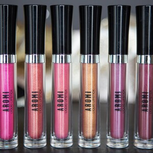 May include: Six clear tubes of liquid lipstick with black caps and silver bands. The tubes are labeled "AROMI LIQUID LIPSTICK". The lipsticks are in shades of pink, red, and gold.