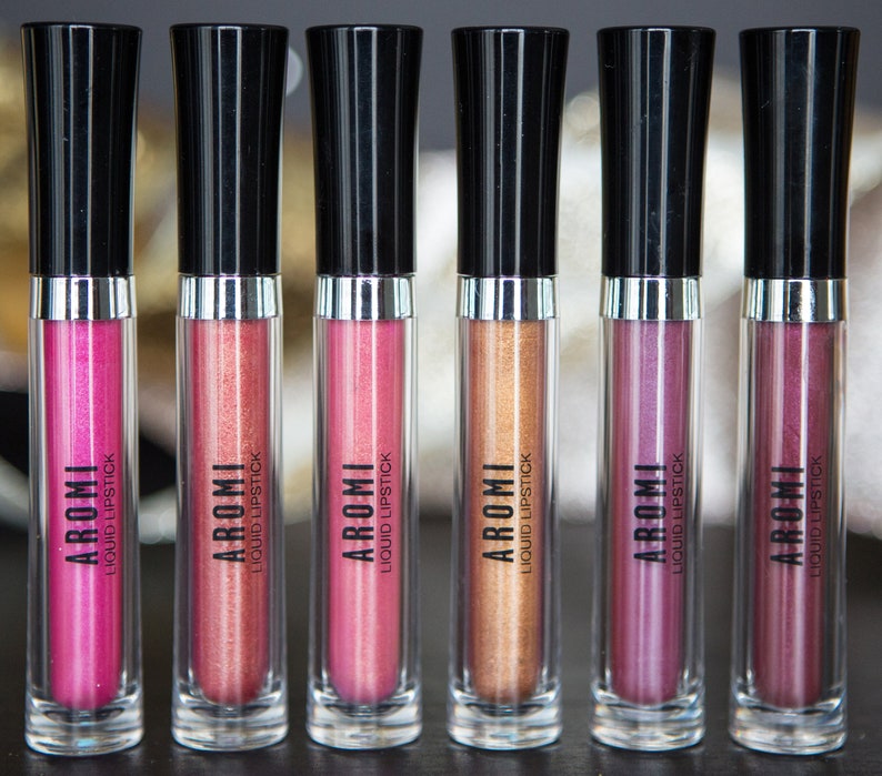 May include: Six clear tubes of liquid lipstick with black caps and silver bands. The tubes are labeled "AROMI LIQUID LIPSTICK". The lipsticks are in shades of pink, red, and gold.