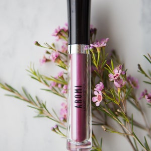 May include: A clear tube of purple liquid lipstick with a black cap and the word "AROMI" printed on the tube. The tube is surrounded by pink flowers.