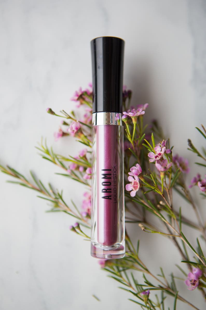 May include: A clear tube of purple liquid lipstick with a black cap and the word "AROMI" printed on the tube. The tube is surrounded by pink flowers.
