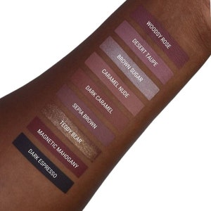 May include: A close-up of a hand with swatches of eyeshadow in various shades of brown, pink, and purple. The swatches are labelled with the names "Woodsy Rose", "Desert Taupe", "Brown Sugar", "Caramel Nude", "Dark Caramel", "Sepia Brown", "Teddy Bear", "Magnetic Mahogany", and "Dark Espresso".