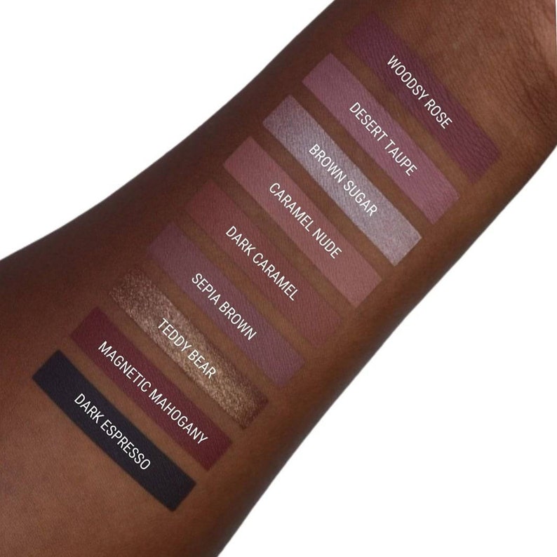 May include: A close-up of a hand with swatches of eyeshadow in various shades of brown, pink, and purple. The swatches are labelled with the names "Woodsy Rose", "Desert Taupe", "Brown Sugar", "Caramel Nude", "Dark Caramel", "Sepia Brown", "Teddy Bear", "Magnetic Mahogany", and "Dark Espresso".