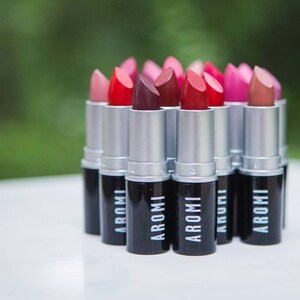 K&ouml;nnte beinhalten: Eine Sammlung von Lippenstift-Tuben in verschiedenen Rot-, Rosa- und Braunt&ouml;nen. Die Tuben sind silberfarben mit schwarzen Kappen und dem Wort "AROMI" auf den Kappen.