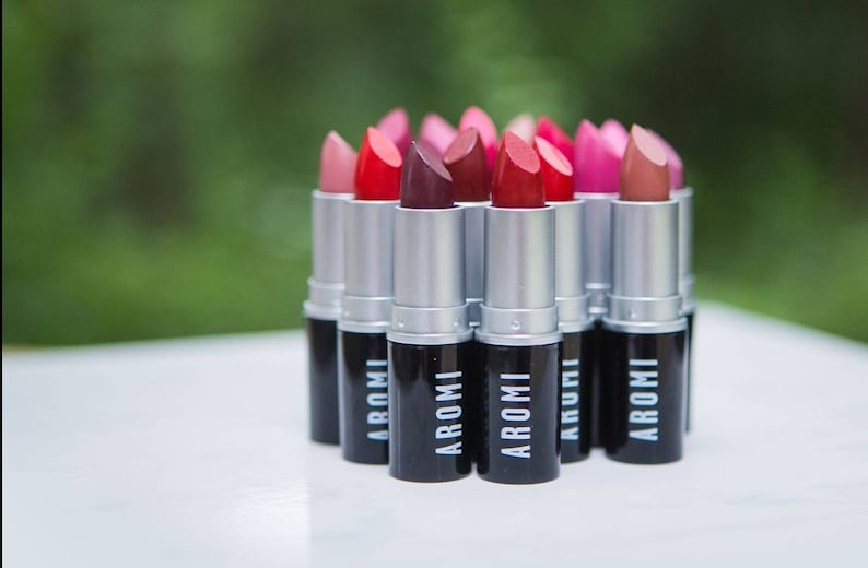K&ouml;nnte beinhalten: Eine Sammlung von Lippenstift-Tuben in verschiedenen Rot-, Rosa- und Braunt&ouml;nen. Die Tuben sind silberfarben mit schwarzen Kappen und dem Wort "AROMI" auf den Kappen.
