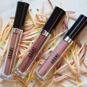 May include: Three tubes of liquid lipstick in a mauve shade. The tubes are black with silver accents and the brand name "AROMI" is printed on the front. The tubes are lying on a surface covered in flower petals.