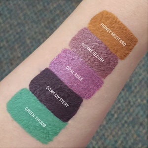 Puede incluir: Muestra de cinco labiales l&iacute;quidos diferentes en el brazo de una persona. Los colores son verde, morado oscuro, rosa, morado claro y naranja. Los nombres de los colores son "Green Thumb", "Dark Mystery", "Opal Rose", "Alpine Bloom" y "Honey Mustard".