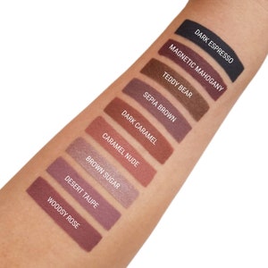 May include: A close-up of a hand with swatches of eyeshadow in various shades of brown, from dark espresso to woodsy rose. The names of each shade are labelled below the swatches.