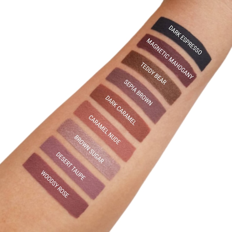 May include: A close-up of a hand with swatches of eyeshadow in various shades of brown, from dark espresso to woodsy rose. The names of each shade are labelled below the swatches.
