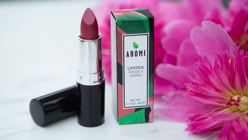 May include: A black lipstick tube with a pink lipstick bullet. The lipstick is in a box with the brand name "AROMI" and the text "LIPSTICK ROUGE A LEVRES" and "NET WT. 0.14 OZ / 4.0 G".