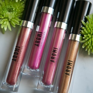 K&ouml;nnte beinhalten: Vier Tuben mit fl&uuml;ssigem Lippenstift in verschiedenen Metallic-Farbt&ouml;nen: Ros&eacute;gold, Pink, Lila und Gold. Jede Tube tr&auml;gt den Markennamen "AROMI" und die Worte "LIQUID LIPSTICK" auf dem Etikett.