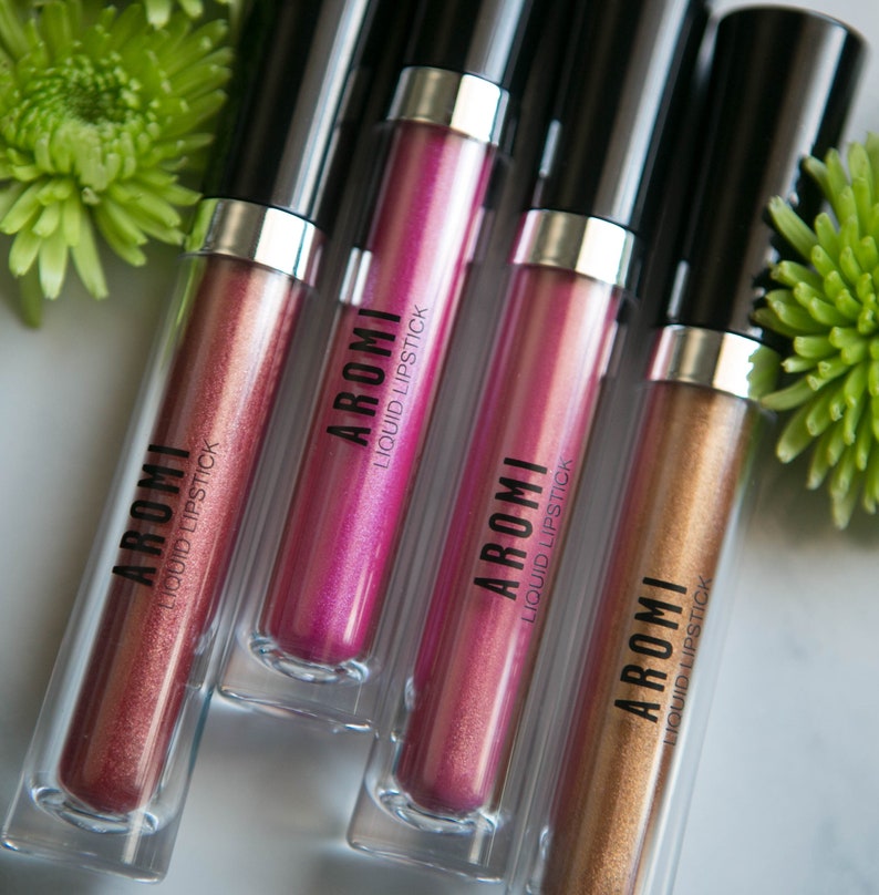 K&ouml;nnte beinhalten: Vier Tuben mit fl&uuml;ssigem Lippenstift in verschiedenen Metallic-Farbt&ouml;nen: Ros&eacute;gold, Pink, Lila und Gold. Jede Tube tr&auml;gt den Markennamen "AROMI" und die Worte "LIQUID LIPSTICK" auf dem Etikett.