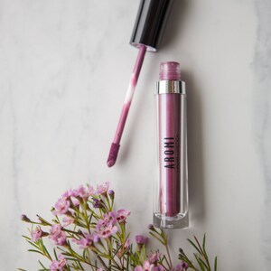 May include: A clear tube of purple liquid lipstick with a black applicator. The tube has the word "AROMI" and "LIQUID LIPSTICK" printed on it. The lipstick is next to a small bouquet of pink flowers.