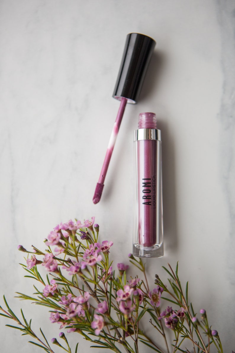 May include: A clear tube of purple liquid lipstick with a black applicator. The tube has the word "AROMI" and "LIQUID LIPSTICK" printed on it. The lipstick is next to a small bouquet of pink flowers.