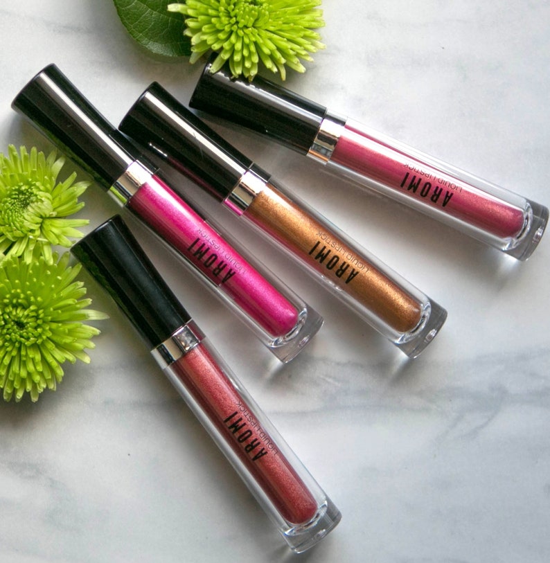K&ouml;nnte beinhalten: Vier Tuben mit fl&uuml;ssigem Lippenstift in verschiedenen Metallic-Farbt&ouml;nen: Pink, Gold, Kupfer und Ros&eacute;gold. Jede Tube hat einen schwarzen Deckel und einen durchsichtigen K&ouml;rper, auf dem der Markenname "AROMI" aufgedruckt ist.