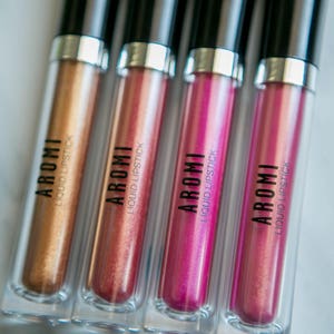 K&ouml;nnte beinhalten: Vier Tuben mit fl&uuml;ssigem Lippenstift in verschiedenen Farbt&ouml;nen von Gold, Rosa und Magenta. Jede Tube hat einen schwarzen Deckel und einen transparenten K&ouml;rper mit dem Wort "AROMI" und "LIQUID LIPSTICK" auf dem Etikett.
