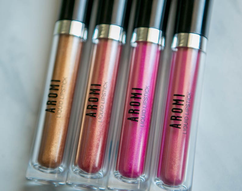 K&ouml;nnte beinhalten: Vier Tuben mit fl&uuml;ssigem Lippenstift in verschiedenen Farbt&ouml;nen von Gold, Rosa und Magenta. Jede Tube hat einen schwarzen Deckel und einen transparenten K&ouml;rper mit dem Wort "AROMI" und "LIQUID LIPSTICK" auf dem Etikett.