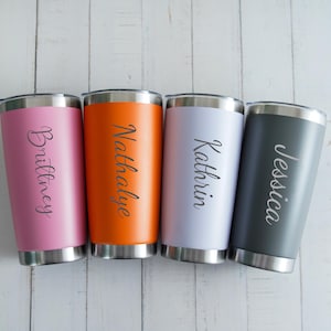Puede incluir: Cuatro vasos personalizados en rosa, naranja, blanco y gris, cada uno con un nombre en escritura elegante. Los vasos de acero inoxidable tienen bordes plateados y tapas negras.