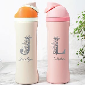 May include: Two personalized water bottles. One is cream-colored with an orange lid, the other is pink with a pink lid. Each bottle has a floral design with a letter and a name: "Jocelyn" and "Linda".
