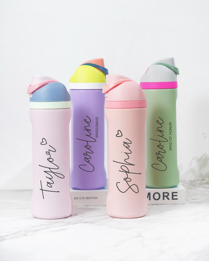 May include: Four personalized water bottles in pastel colors: pink, purple, and green. Each bottle has a different name in cursive script, such as "Taylor" and "Sophia", and a heart symbol. The lids are multi-colored.