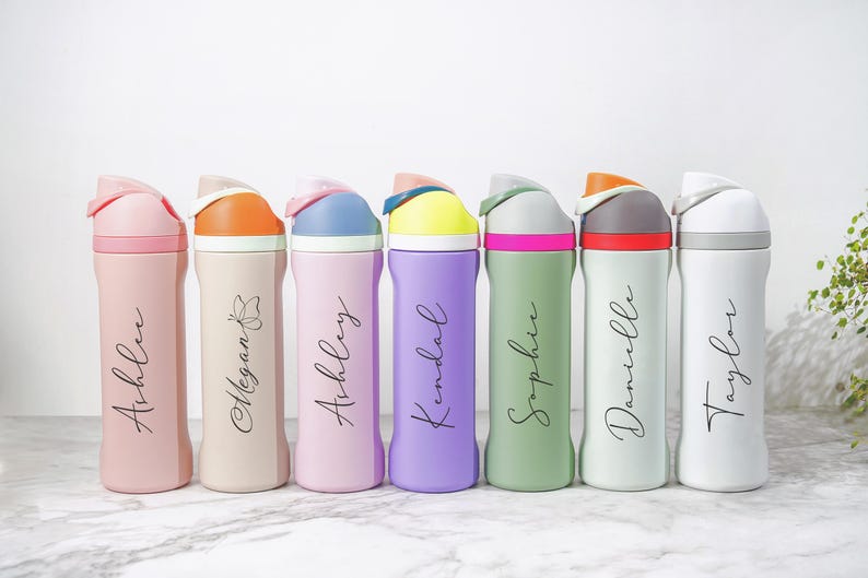 May include: A collection of seven colorful water bottles with flip-top lids. Each bottle is a different pastel color, including pink, beige, purple, green, and white. The bottles are personalized with names in a cursive font.
