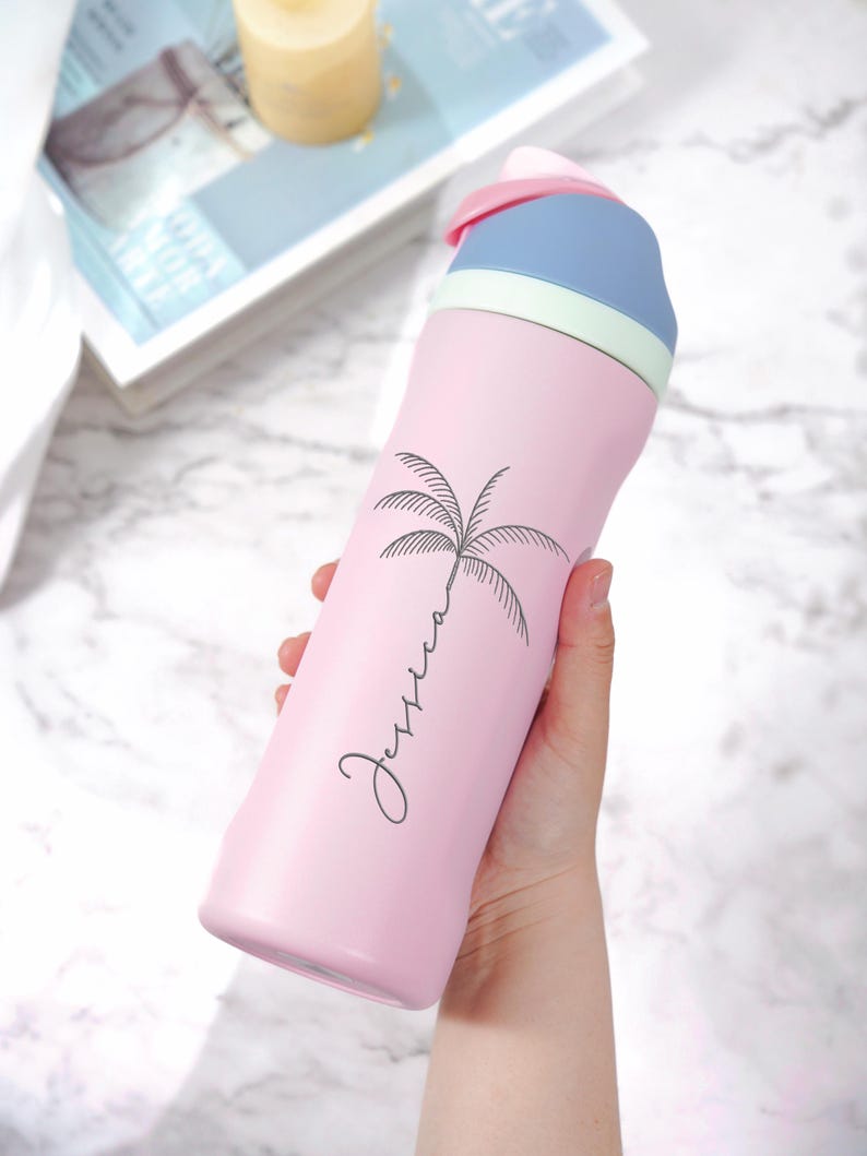 May include: A pink insulated water bottle with a black palm tree design and the name "Jessica" in cursive. The bottle has a blue and pink lid with a light green accent. The bottle is held by a person.