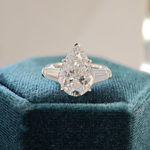 Puede incluir: Un anillo de tono plateado con una gran piedra central en talla pera sujeta por garras, flanqueada por dos piedras laterales en talla baguette cónica. La banda de metal es pulida y lisa, mostrando un diseño clásico de tres piedras.