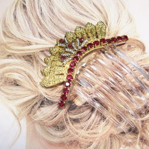 May include: A gold-toned hair comb with a decorative design featuring red and clear gemstones. The comb is adorned with a fan-like embellishment and a row of red stones. The clear plastic comb teeth are visible.
