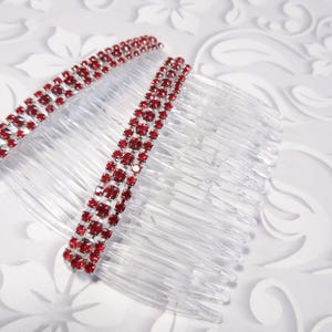 May include: Two clear plastic hair combs with a row of red rhinestones. The combs are on a white background with a floral pattern. The rhinestones are set in a silver-colored metal.