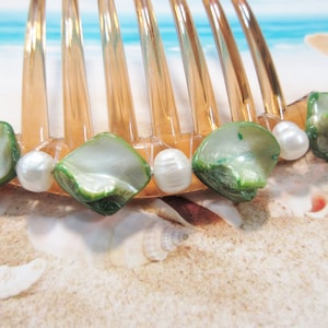 May include: A clear, amber-colored hair comb adorned with green and white iridescent shells and white pearls. The comb's teeth are evenly spaced, and the shells and pearls are attached to the base, creating a beachy, ocean-inspired accessory.