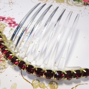 May include: A clear plastic hair comb with a row of dark red rhinestones attached to the bottom. The rhinestones are set in gold.