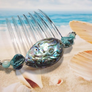 May include: A clear plastic hair comb with a large iridescent abalone shell and turquoise blue glass beads. The comb is lying on a sandy beach with seashells.