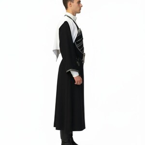 May include: A person in a traditional black and white outfit, viewed from the side. The outfit features a long black coat, a white shirt, and a white shawl. The coat has silver trim, and the person is wearing black boots.