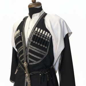 May include: A traditional black and white outfit with silver detailing, presented on a mannequin. The outfit features a long coat, a vest with cartridge holders, and a decorative dagger. A white shirt and cape complete the ensemble.