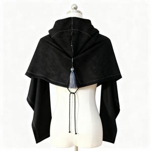 Puede incluir: Una capa con capucha negra con un diseño de capa, exhibida en un maniquí. La capa presenta una capucha con costuras visibles, una borla decorativa y mangas largas. El diseño incluye un cierre de lazo en la parte delantera.