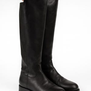 May include: A pair of tall, black leather boots. The boots have a smooth texture and a classic design, with a flat sole and a knee-high shaft. The boots are displayed against a white background.