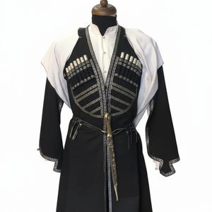 May include: A traditional black and white outfit with silver detailing, displayed on a mannequin. The outfit includes a long coat, a vest with decorative silver detailing, and a matching belt with a decorative knife.