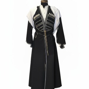 May include: A traditional black and white garment, possibly a coat or robe, displayed on a mannequin. The garment features intricate silver-coloured embellishments and a decorative belt with a knife. The sleeves are long, and the garment has a high collar.