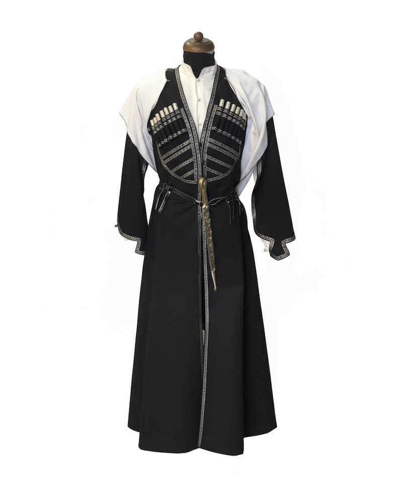 May include: A traditional black and white garment, possibly a coat or robe, displayed on a mannequin. The garment features intricate silver-coloured embellishments and a decorative belt with a knife. The sleeves are long, and the garment has a high collar.