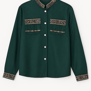 Puede incluir: Camisa verde oscuro de manga larga, con detalles bordados geométricos dorados, marrones y rojos en el cuello, puños y bolsillos. Tiene dos bolsillos delanteros y botones blancos. La camisa está colgada de una percha negra.