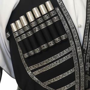 May include: Close-up of a black vest with silver decorative trim and seven silver-topped cylindrical holders. The vest is worn over a white shirt. The vest has horizontal silver stripes and a decorative silver border.