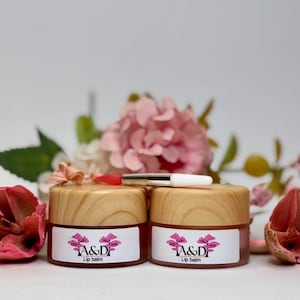 Puede incluir: Dos pequeños frascos de madera con bálsamo labial rosa en su interior. Los frascos tienen etiquetas blancas con el texto "A&D Lip balm" y un diseño floral rosa. Los frascos están sobre una superficie blanca con flores rosas.