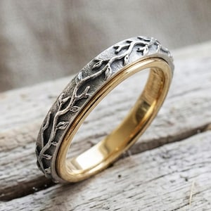 May include: A two-tone ring featuring a polished gold-coloured inner band and a textured, oxidised silver-coloured outer band. The outer surface displays a detailed, raised relief pattern of delicate, winding branches with small leaves.