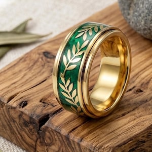 May include: A gold-colored ring with a green band featuring a leaf design. The ring is set on a wooden surface with dried leaves and a gray stone in the background. The ring's design is a repeating pattern of leaves.