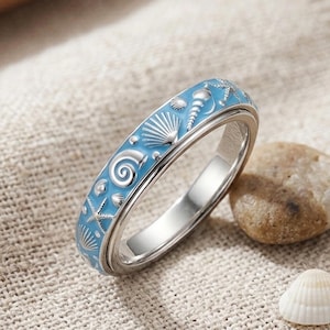May include: A silver-toned metal ring featuring a light blue enamel inlay decorated with various embossed seashell and starfish motifs. The band has a smooth, polished interior surface and a detailed, textured outer surface with nautical elements.